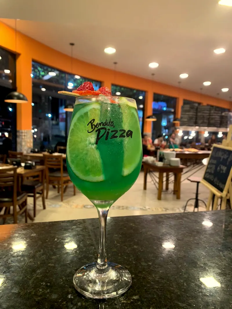 Drink verde na taça com a logo da Bendita Pizza, diante da parede laranja do salão