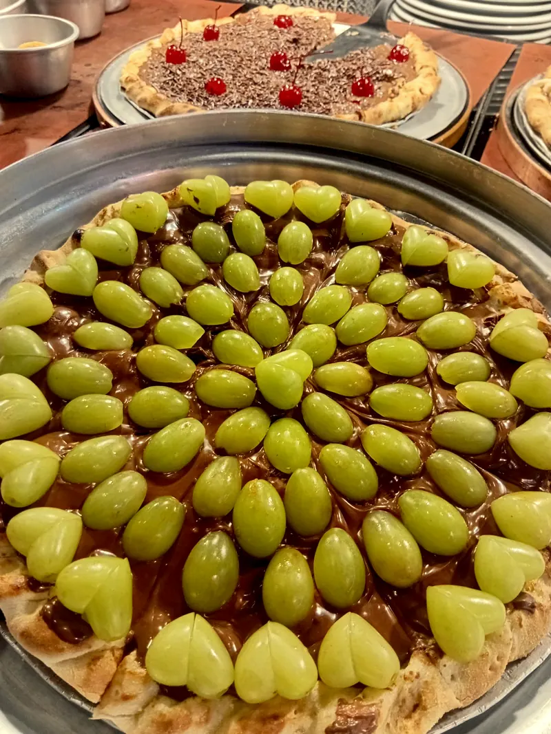 Pizza doce de chocolate com uvas verdes, e outra com cerejas atrás